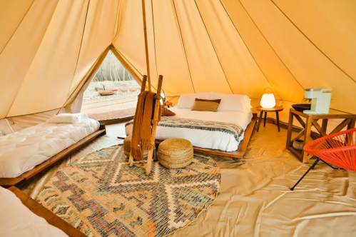 Cozy glamping tent interior with two beds, a rug, and warm lighting, surrounded by nature.